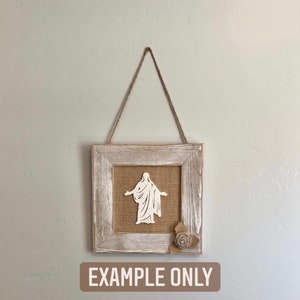 May include: A white silhouette of a person with outstretched arms is framed in a rustic whitewashed wood frame with a burlap background. A burlap flower is in the bottom right corner of the frame. The frame is hanging from a twine rope. The text "EXAMPLE ONLY" is printed below the frame.