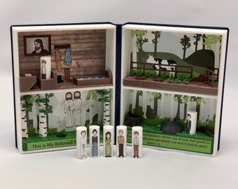 Puede incluir: Un libro de madera en miniatura con una escena emergente que representa a una familia en un entorno de bosque. El libro está abierto y la escena emergente incluye una vaca, árboles y una familia de cinco. El libro tiene el texto "This is My Beloved S" y "lack wisdom, let him ask of God, that giveth to all, and upbraideth not, and it shall be given James 1:5" impreso en las portadas delantera y trasera.