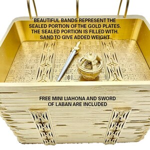 May include: A gold-coloured, rectangular box with a decorative design. The box is filled with sand and contains a small sword and a round object. The text on the box reads: "BEAUTIFUL BANDS REPRESENT THE SEALED PORTION OF THE GOLD PLATES. THE SEALED PORTION IS FILLED WITH. SAND TO GIVE ADDED WEIGHT FREE MINI LIAHONA AND SWORD OF LABAN ARE INCLUDED"