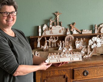 May include: A woman holding a wooden religious diorama depicting scenes from the Bible. The diorama is made of layered wood and features figures and structures in a natural wood and white color scheme. The scenes include the crucifixion and the last supper.