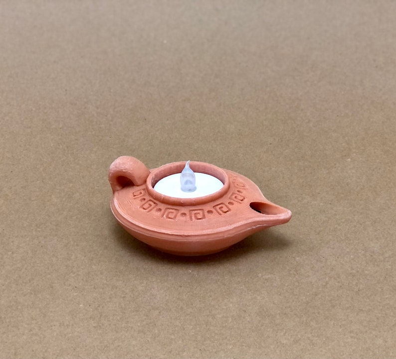 Puede incluir: Una l&aacute;mpara de aceite de terracota con una vela LED blanca en su interior. La l&aacute;mpara tiene un dise&ntilde;o decorativo alrededor del borde.