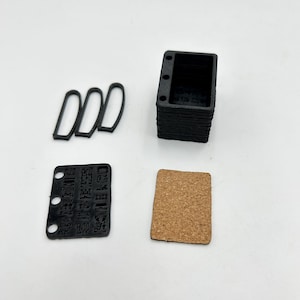 Puede incluir: Un conjunto de piezas impresas en 3D de color negro para un proyecto de bricolaje. El conjunto incluye una caja, tres juntas de goma, un pequeño trozo de corcho y una etiqueta de plástico negro con el texto "MADE BY RECYCLED PLASTIC DIY & RE-USE PIECES".