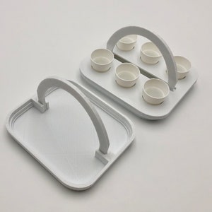 May include: White plastic tray with a handle, divided into six sections, each holding a small white cup. The tray is designed to hold six small cups.
