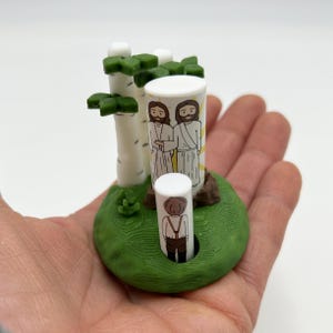 May include: A 3D printed scene with two figures standing in front of a birch tree. The figures are white with black hair and are wearing white robes. The birch tree is white with green leaves. The scene is set on a green base with a brown log.