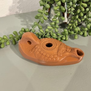 May include: A terracotta-colored oil lamp with a detailed design. The lamp features a central circular opening, a spout, and decorative patterns. The lamp is a warm, earthy tone, and is a decorative item.