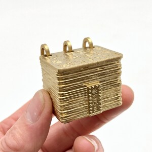 May include: A small, gold-colored, rectangular pendant with three loops on top. The pendant has etched details and a small door with a sun, moon, and star design. The pendant is held in a hand.