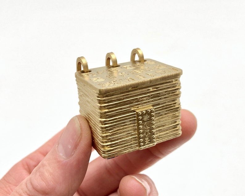 May include: A small, gold-colored, rectangular pendant with three loops on top. The pendant has etched details and a small door with a sun, moon, and star design. The pendant is held in a hand.
