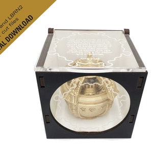 May include: A gold-colored sphere with intricate details sits inside a black wooden box with a clear acrylic lid. The lid features a white inscription that reads "We follow their obuna of sorrow into a far Desper land of promise, Aims 37,45".
