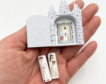 May include: A white miniature model of a church with a small picture of Jesus inside. The church is open and there are two small figures of a bride and groom standing in front of it.