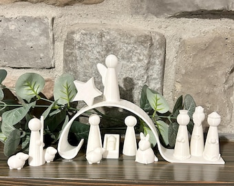 May include: A white nativity scene set with a star, angel, and various figurines, including sheep, wise men, and a manger. The figures are arranged on a wooden surface with a brick background and greenery.