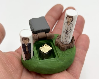 Puede incluir: Un modelo 3D impreso de una escena de la película 'La Princesa Prometida'. El modelo presenta dos figuras, un ladrillo de oro y una base verde. Las figuras están impresas en cilindros blancos y representan personajes de la película. El ladrillo de oro se coloca en una pequeña abertura en la base.
