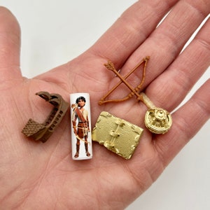 May include: A set of five small, brown and gold plastic toys. The toys include a miniature ship, a character holding a bow and arrow, a bow and arrow, a book, and a round object with a design.