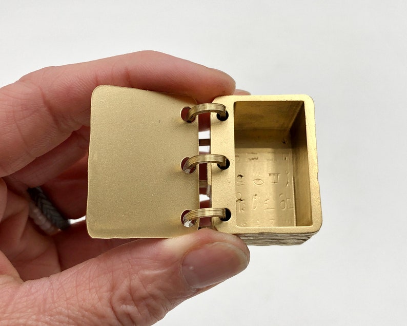 May include: A small, open, brass-colored book with three metal rings. The pages are rectangular and the interior has etched symbols. The book is held open, revealing the inside. The book is a unique, handcrafted item.