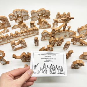 May include: A collection of tan-colored, 3D-printed religious figures and structures, including trees, people, and crosses. A card with the text "THE WEEK THAT LED TO EASTER" is held in the foreground. The scene depicts the Palm Sunday entry into Jerusalem.