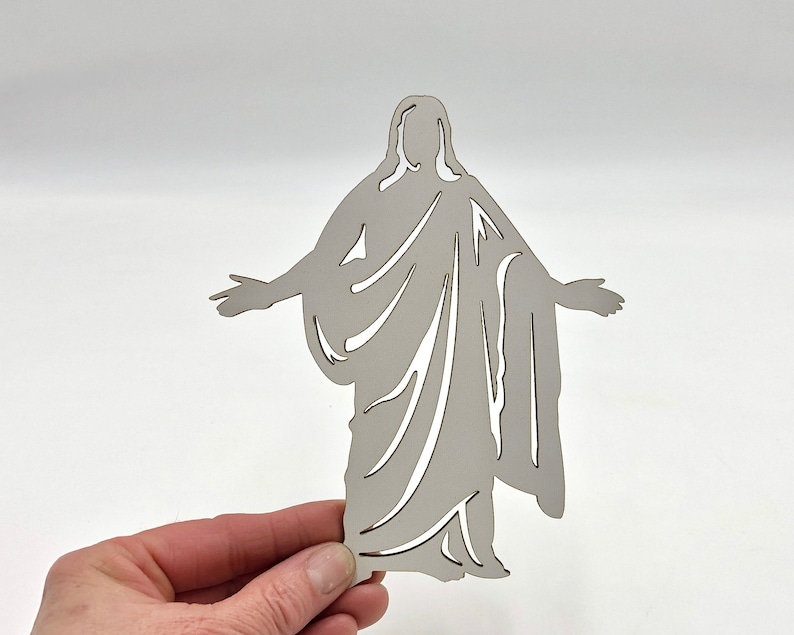 May include: A white cutout of a person with arms outstretched, resembling a religious figure. The cutout is made of wood and has a simple, elegant design.