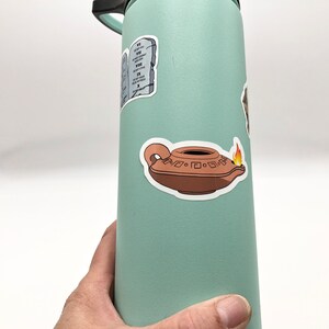 May include: A light green water bottle with a sticker of a clay oil lamp with a flame on it. The sticker is on the right side of the bottle. There is also a sticker of a stone tablet with Roman numerals on it.