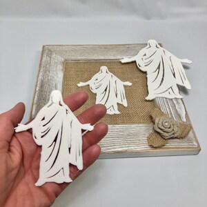 May include: Three white cutout figures of a person in a robe with arms outstretched. The figures are displayed on a burlap background in a wooden frame.
