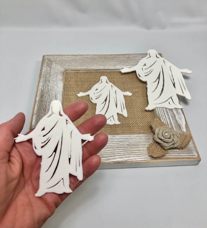 May include: Three white cutout figures of a person in a robe with arms outstretched. The figures are displayed on a burlap background in a wooden frame.
