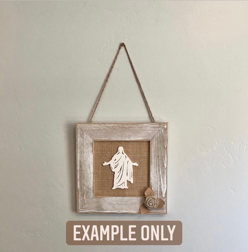 May include: A white silhouette of a person with outstretched arms is framed in a rustic whitewashed wood frame with a burlap background. A burlap flower is in the bottom right corner of the frame. The frame is hanging from a twine rope. The text "EXAMPLE ONLY" is printed below the frame.