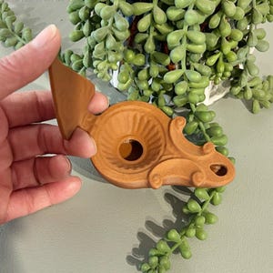 May include: A decorative, terracotta-colored object with an ornate design. The object has a circular base with a central hole and a stylized arrow-shaped element. The design includes scroll-like details and a textured surface.