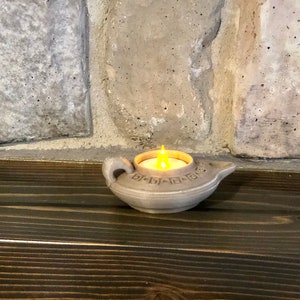 Puede incluir: Una peque&ntilde;a l&aacute;mpara de aceite de cer&aacute;mica gris con una vela en su interior. La l&aacute;mpara tiene un dise&ntilde;o decorativo alrededor del borde y un pico en un lado. La vela est&aacute; encendida y proyecta un c&aacute;lido resplandor.