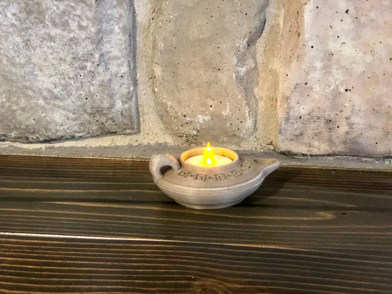Puede incluir: Una peque&ntilde;a l&aacute;mpara de aceite de cer&aacute;mica gris con una vela en su interior. La l&aacute;mpara tiene un dise&ntilde;o decorativo alrededor del borde y un pico en un lado. La vela est&aacute; encendida y proyecta un c&aacute;lido resplandor.