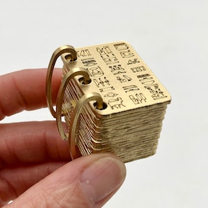 May include: A small, gold-colored metal book with engraved hieroglyphs. The book is held together by three curved metal rings. The pages of the book are visible, and the overall design suggests a miniature replica of an ancient artifact.