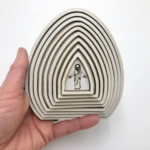 May include: White wooden cutout of a figure with outstretched arms, surrounded by concentric circles, resembling a sunburst or a halo.