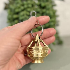 May include: A gold-colored keychain featuring a detailed, ornate lantern design. The lantern has a decorative top and base, with intricate patterns and cutouts. The keychain is attached to a silver-colored split ring and clasp.