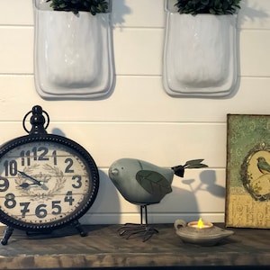 Puede incluir: Un reloj negro con n&uacute;meros romanos y una esfera blanca, una figurita de p&aacute;jaro azul, una peque&ntilde;a l&aacute;mpara de aceite y un cuadro enmarcado con un p&aacute;jaro azul en una rama. El cuadro tiene un fondo verde con un marco dorado.