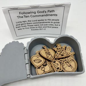 Puede incluir: Una caja gris en forma de corazón que contiene fichas de madera con ilustraciones grabadas y números, que representan los Diez Mandamientos. Un papel con el texto "Following God's Path The Ten Commandments" está encima de la caja.