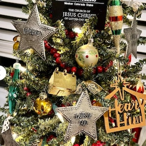 May include: A decorated Christmas tree with various ornaments. Silver star ornaments with the words "REDEEMER" and "LAMB OF GOD" are visible. Other ornaments include gold and red baubles, and a wooden cutout with the words "Hear Him".