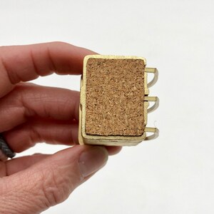 May include: A small, rectangular, gold-colored metal object with a cork insert. The object has three curved metal pieces on one side. The cork is a light brown color. The object is held in a person's hand.