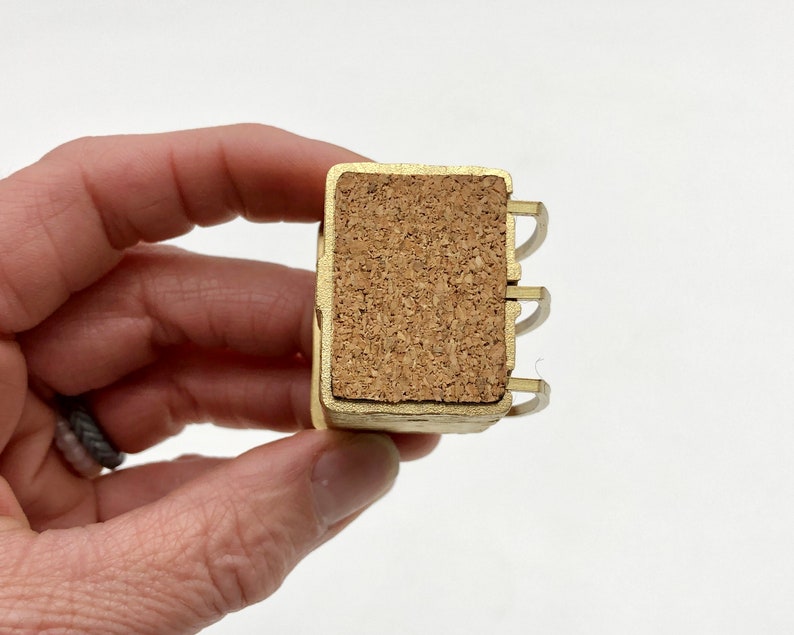 May include: A small, rectangular, gold-colored metal object with a cork insert. The object has three curved metal pieces on one side. The cork is a light brown color. The object is held in a person's hand.