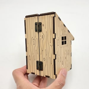 May include: A wooden model of a small cabin with a hinged door and a window. The cabin is made of laser-cut wood and has a wood grain pattern.