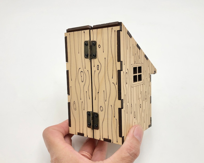 May include: A wooden model of a small cabin with a hinged door and a window. The cabin is made of laser-cut wood and has a wood grain pattern.