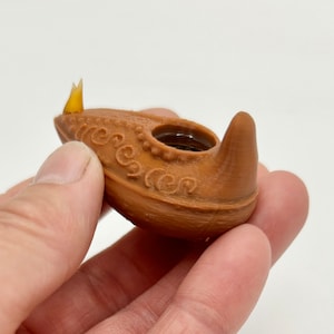 Puede incluir: Una pequeña lámpara de aceite de cerámica marrón con un diseño decorativo. La lámpara tiene un pequeño pico y un porta mechas. La lámpara está en una mano.
