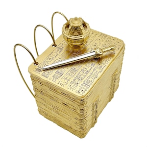 Puede incluir: Un libro de metal dorado con una corona dorada en la parte superior y una espada dorada que sobresale del lado. El libro está cubierto de jeroglíficos.