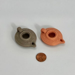 Puede incluir: Dos peque&ntilde;as l&aacute;mparas de aceite de cer&aacute;mica con un dise&ntilde;o de clave griega. Una l&aacute;mpara es gris y la otra es naranja.