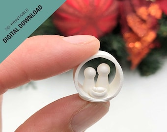 Puede incluir: Adorno de pesebre de Navidad impreso en 3D blanco con un pesebre que contiene un bebé y dos figuras de pie a su lado. El adorno se sostiene en una mano sobre un fondo borroso de rojo y verde.