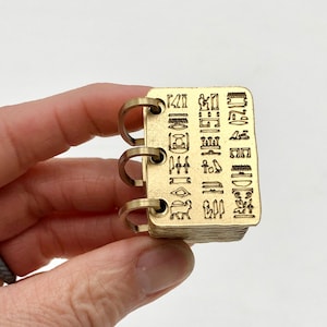 May include: A small, rectangular brass notebook with three metal rings. The cover is engraved with hieroglyphs. The pages are stacked and appear to be made of the same brass material. The notebook is held in a person's hand against a white background.
