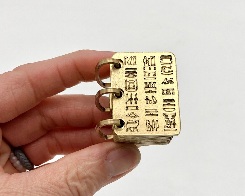May include: A small, rectangular brass notebook with three metal rings. The cover is engraved with hieroglyphs. The pages are stacked and appear to be made of the same brass material. The notebook is held in a person's hand against a white background.