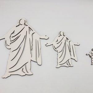 May include: Three white wooden cutout figures of a person in a robe with arms outstretched. The figures are different sizes.