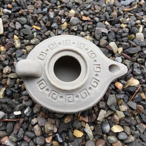 Puede incluir: Una l&aacute;mpara de aceite de cer&aacute;mica gris con un dise&ntilde;o decorativo alrededor del borde. La l&aacute;mpara tiene un cuerpo redondo con un peque&ntilde;o pico y una abertura circular en el centro.
