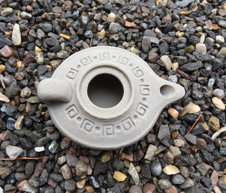 Puede incluir: Una l&aacute;mpara de aceite de cer&aacute;mica gris con un dise&ntilde;o decorativo alrededor del borde. La l&aacute;mpara tiene un cuerpo redondo con un peque&ntilde;o pico y una abertura circular en el centro.