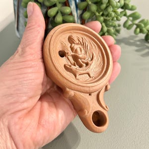 Puede incluir: Una lámpara de aceite de estilo antiguo, de color marrón claro, con un detallado relieve tallado. El relieve representa una figura y un cocodrilo. La lámpara tiene un asa y una boquilla para la mecha.