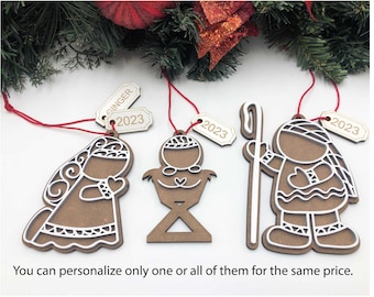 Puede incluir: Cuatro adornos navideños de madera que representan la escena de la Natividad. Los adornos son marrones con detalles blancos e incluyen a María, José, el niño Jesús y un pastor. Cada adorno tiene una etiqueta con el año 2023 impreso en ella. El texto "Puedes personalizar solo uno o todos ellos por el mismo precio." está impreso debajo de los adornos.