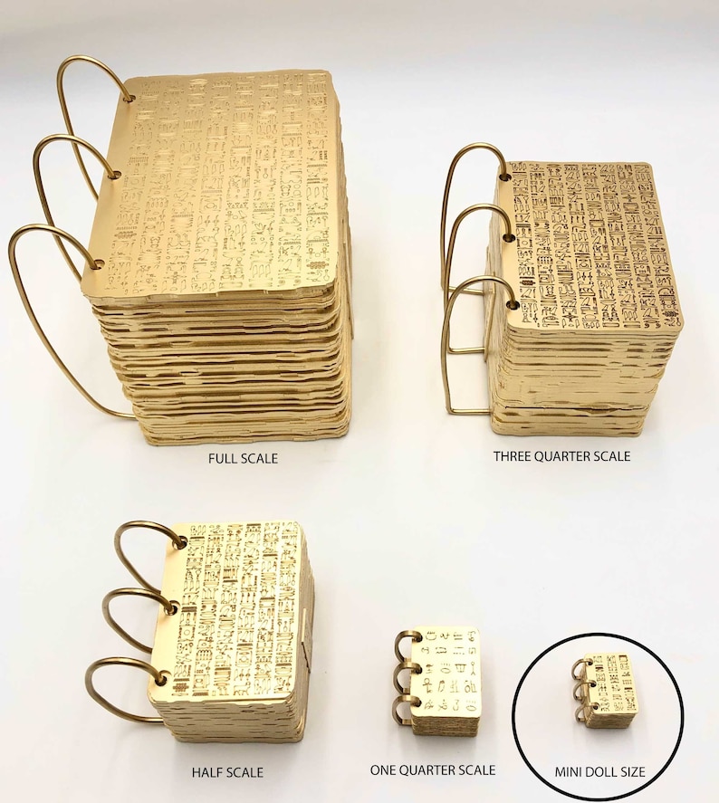 May include: A collection of gold-colored miniature notebooks, each with a unique size and design. The notebooks are bound with gold wire and feature intricate hieroglyphic patterns. Sizes range from full scale to mini doll size.