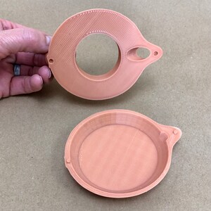 Puede incluir: Dos recipientes redondos de pl&aacute;stico de color melocot&oacute;n impresos en 3D con una peque&ntilde;a manija en el lateral. El recipiente superior tiene una abertura circular en el centro.