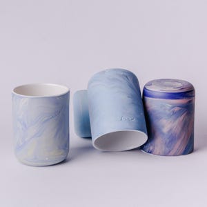 May include: Four ceramic mugs with a marbled glaze in shades of blue, white, and pink. The mugs are stacked on a white surface.